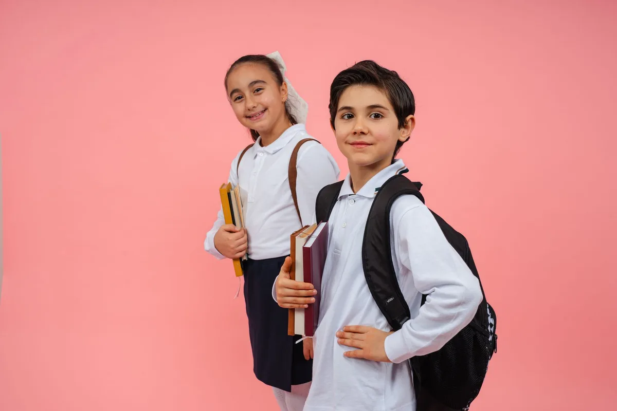 Two children in school uniforms holding books and backpacks against a colored backdrop