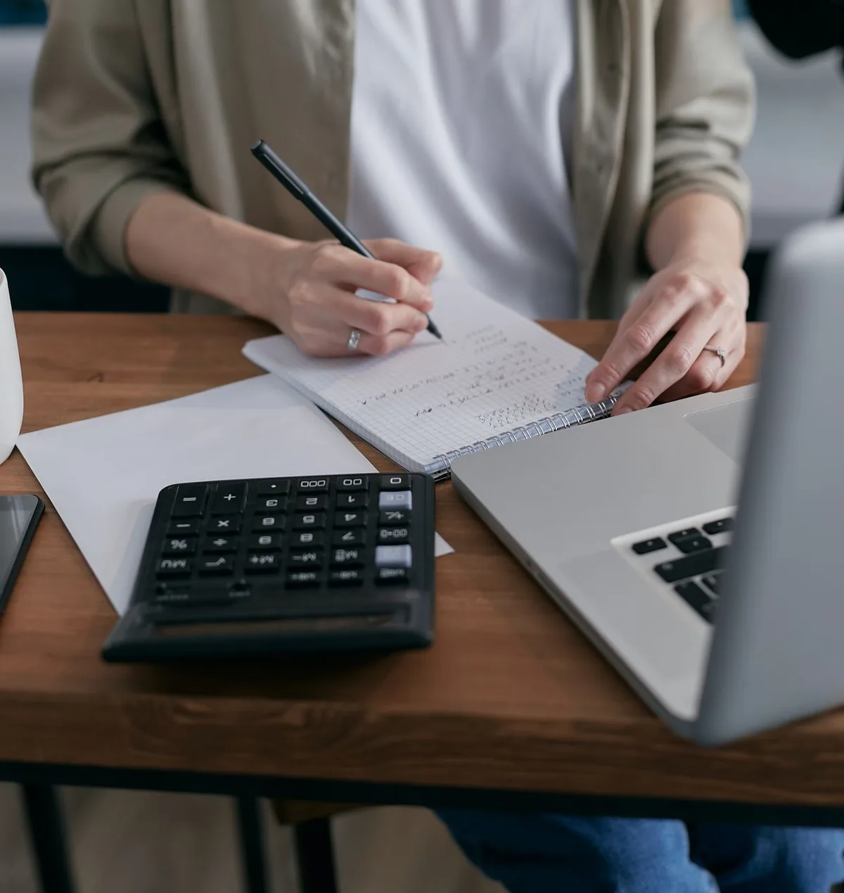 Person calculating finances in notebook with calculator and laptop on wooden desk.