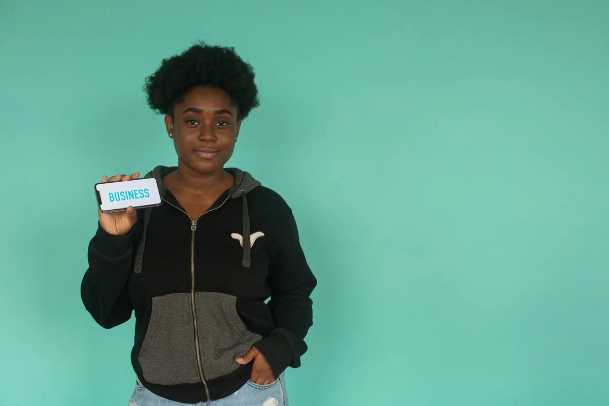 Confident woman in hoodie holding smartphone displaying 'business' against teal background.